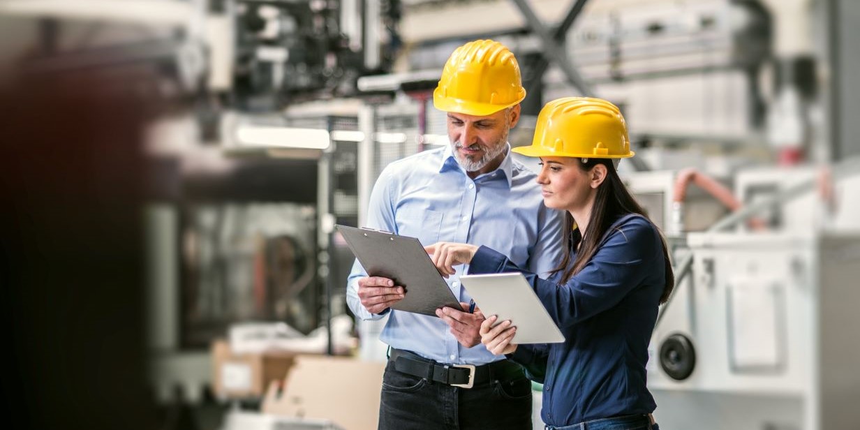 A man and a woman in yellow hard hats look consult a tablet and clipboard in an industrial setting.