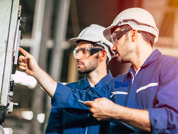 two workers in blue coveralls and white hard hats operating a control panel in an industrial setting