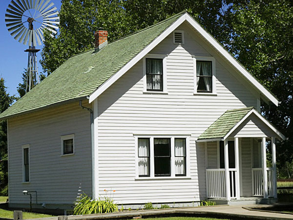 white two-story house with green roof, front porch, and surrounding greenery, with a windmill in the background