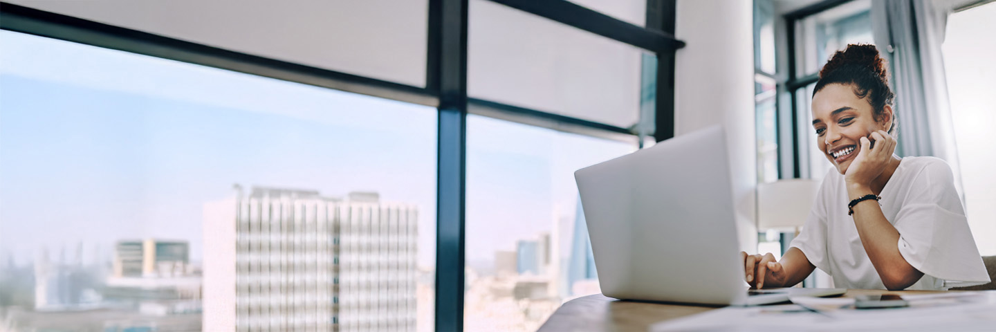 professional working at a desk with laptop, city skyline visible through large windows, desk lamp and documents in view