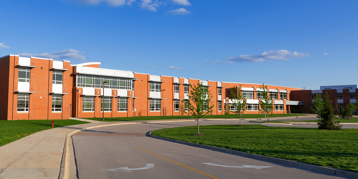 modern brick school building exterior with large windows, landscaped campus grounds, and access road, representing educational facility design and school property safety