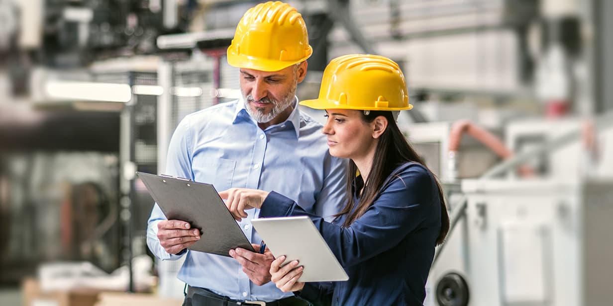 industrial workers in safety gear reviewing documents on clipboard and tablet in a factory setting with machinery in the background