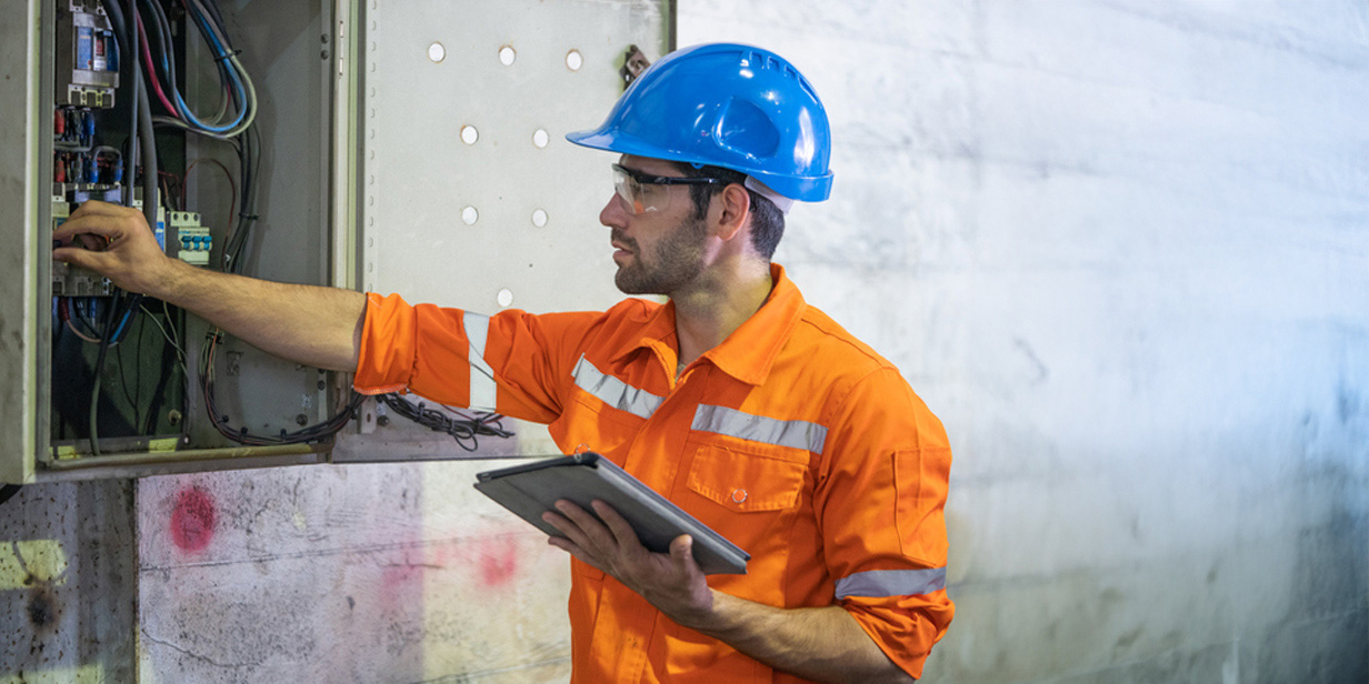 industrial electrician in orange safety uniform and blue hard hat inspecting electrical control panel while using a digital tablet in a factory setting