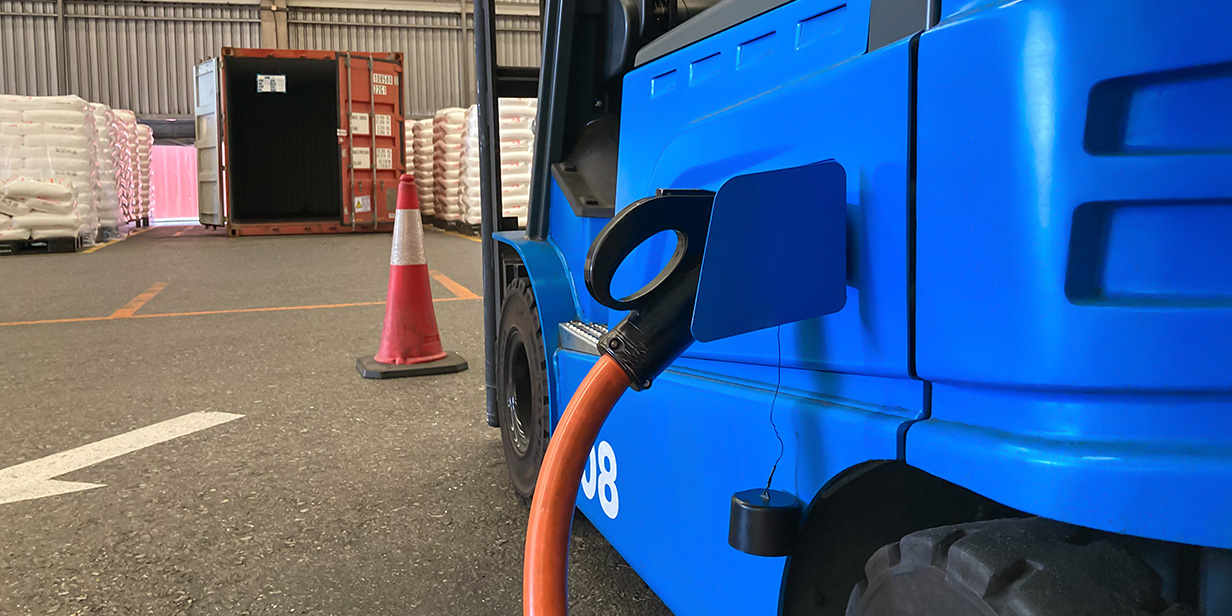 electric forklift charging battery in warehouse with safety cone and stacked industrial materials in background