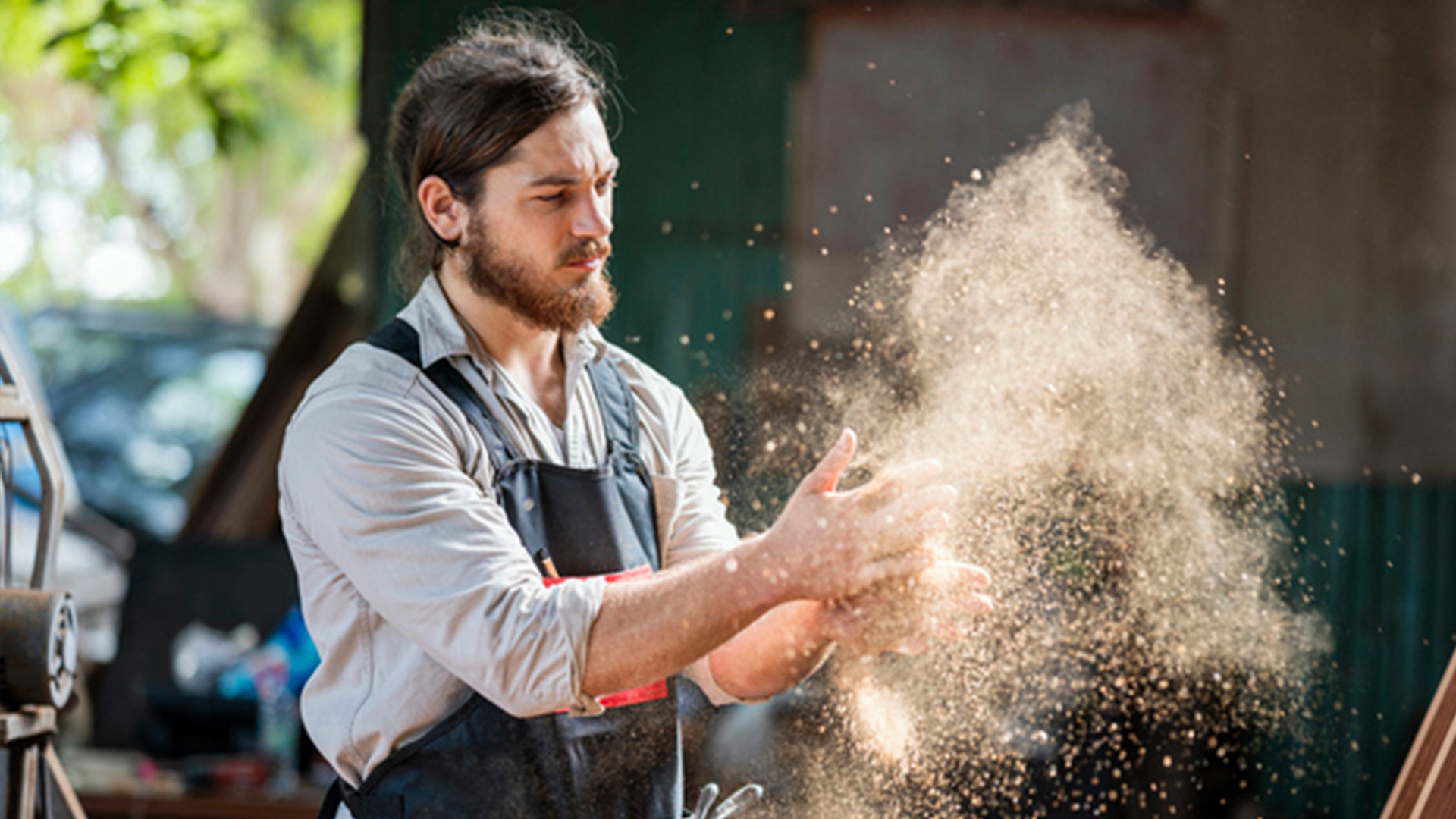 woodworker in a workshop apron clapping hands, creating a cloud of sawdust during woodworking process, highlighting wood dust exposure and safety risks