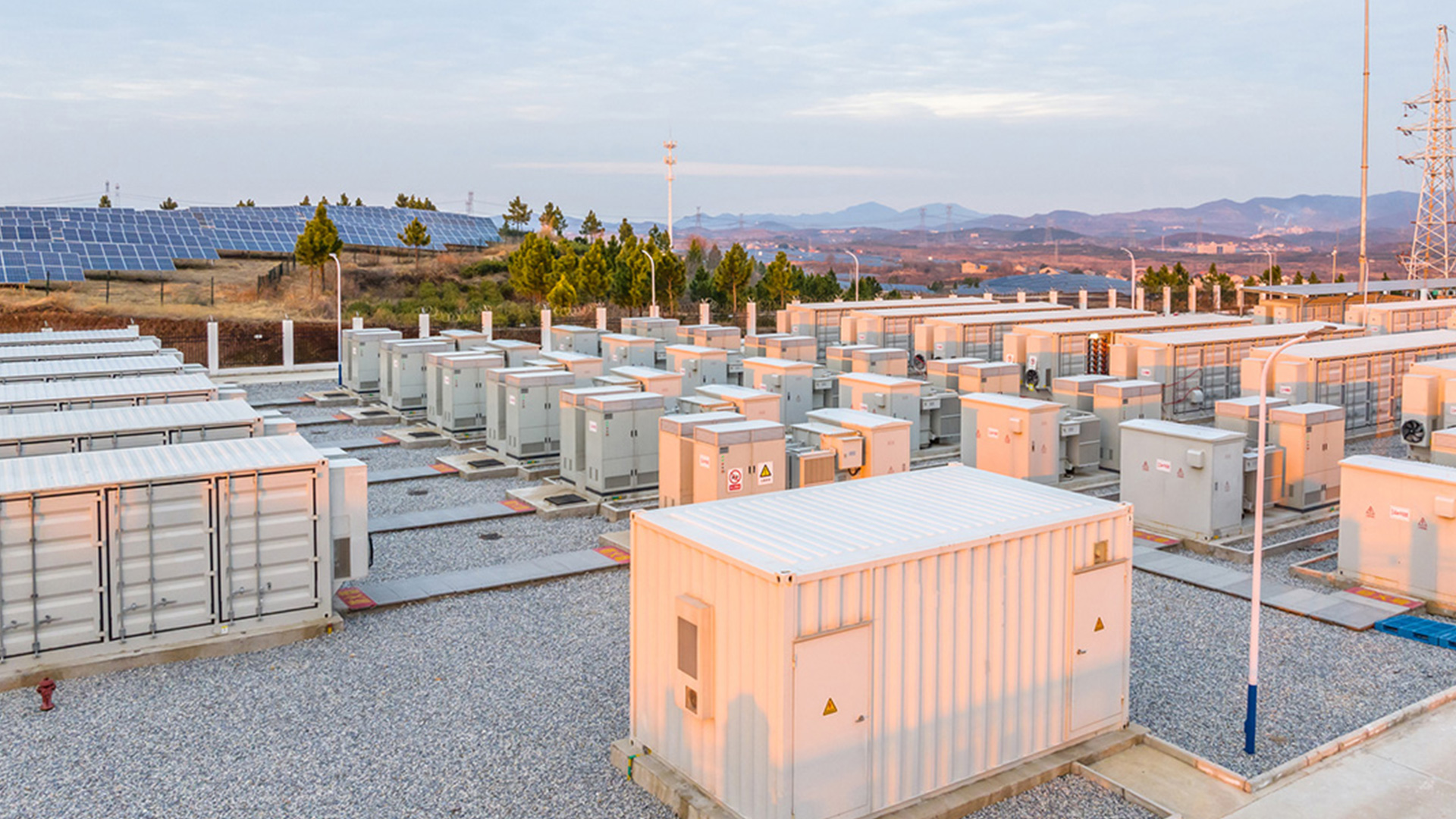 large battery energy storage system (BESS) facility with rows of battery containers and electrical equipment connected to a nearby solar power installation, supporting renewable energy storage and grid reliability