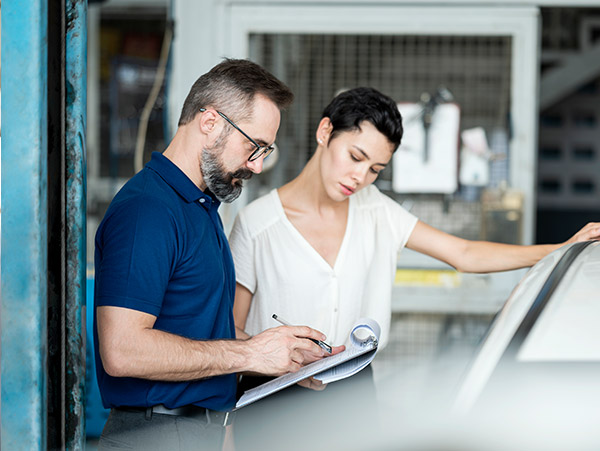 two people next to a car in a garage, one holding a clipboard